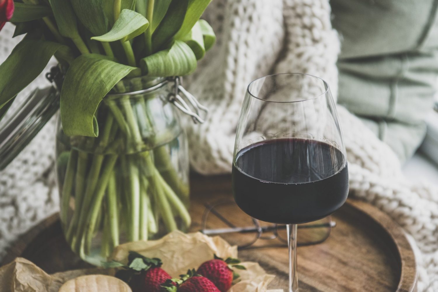 Glass of red wine, snacks and spring red tulips Wine snack set with flowers. Glass of red wine, cheese, roasted almonds, strawberries and bouquet of Spring red tulips on wooden tray over white knitted blanket, close-up. Relaxed mood concept